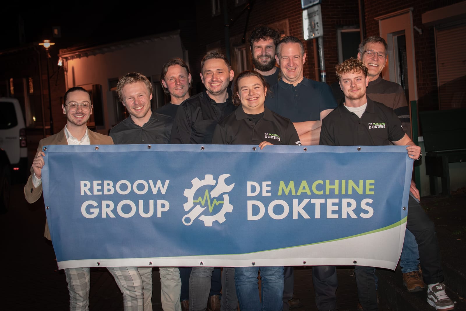 Het team houdt een banner vast met REBOOW GROUP en DE MACHINEDOKTERS erop.