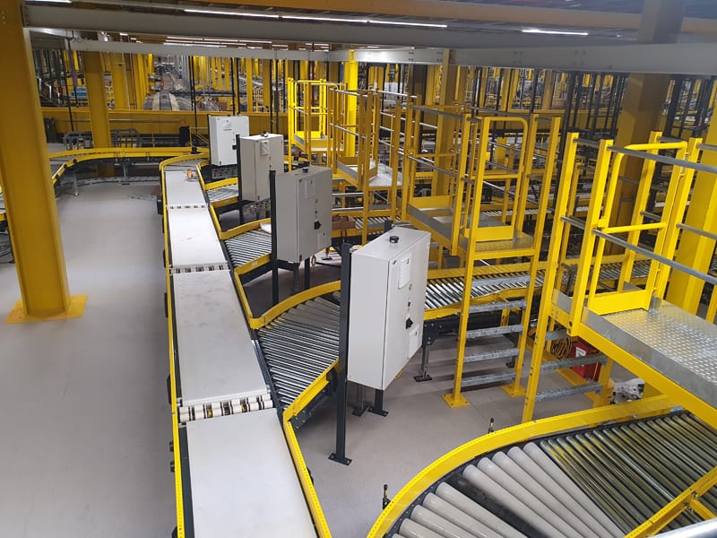 Industrieel transportsysteem met rollenbanen en gele leuningen in een fabriekshal.